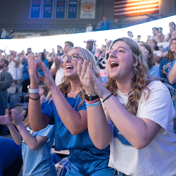 Two female sycamore fans cheer and clap and are surrounded by other cheering students in the fan section of a basketball game. 